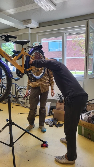 Zwei Personen arbeiten gemeinsam an einem gelb-blauen Kinderfahrrad, das in einem Montageständer eingespannt ist.